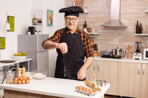 Senior baker sprinkling whear flour on home kitche table for tasty recipe. Retired chef with bonete and apron, in kitchen uniform sprinkling sieving sifting ingredients by hand. photo