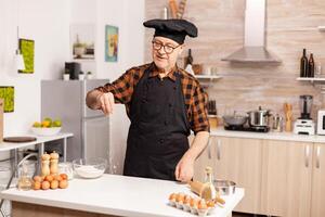 Experienced baker spreading flour in home kitchen for food preparation. Retired senior chef with bonete and apron, in kitchen uniform sprinkling sieving sifting ingredients by hand. photo