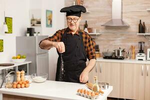 Elderly man preparing food spreading flour in home kitchen for food preparation. Retired senior chef with bonete and apron, in kitchen uniform sprinkling sieving sifting ingredients by hand. photo