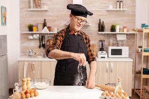 Smiling senior baker in home kitchen using bio ingredients for tasty recipe. Retired chef with bonete and apron, in kitchen uniform sprinkling sieving sifting ingredients by hand. photo