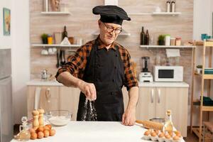 Chef wearing bonete and apron while preparing ingredient for delicious recipe in home kitchen. Cook in kitchen uniform sprinkling sieving sifting ingredients by hand. photo