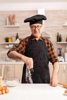 Happy retired chef preparing homemade pizza using bio wheat flour. Retired senior chef with bonete and apron, in kitchen uniform sprinkling sieving sifting ingredients by hand. photo