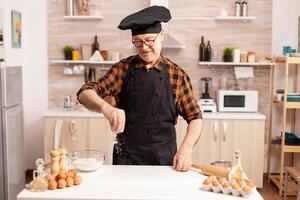 Senior chef preparing homemade bread sprinkling wheat flour on kitchen table. Retired senior chef with bonete and apron, in kitchen uniform sprinkling sieving sifting ingredients by hand. photo