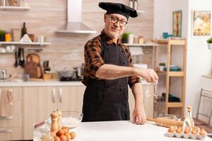 Senior man wearing apron smiling while sprinkling what flour with hand on kitchen table. Retired chef with bonete and apron, in kitchen uniform sprinkling sieving sifting ingredients by hand. photo