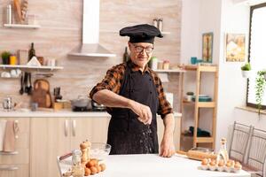 Senior baker in home kitchen during preparation of cookies using wheat flour. Retired senior chef with bonete and apron, in kitchen uniform sprinkling sieving sifting ingredients by hand photo