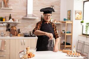 Senior baker preparing bread using bio wheat flour in home kitchen wearing apron and bonete. Cook in kitchen uniform sprinkling sieving sifting ingredients by hand. photo