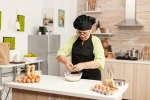 mujer rompe huevo encima harina haciendo masa para panadería productos mayor Pastelería cocinero agrietamiento huevo en vaso cuenco para pastel receta en cocina, mezcla por mano, amasadura ingredientes preparando hecho en casa pastel foto