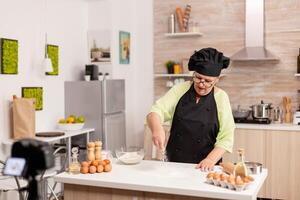 Senior woman creating content for culinary blog preparing tasty bread. Retired blogger chef influencer using internet technology communicating, shooting blogging on social media with digital equipment. photo