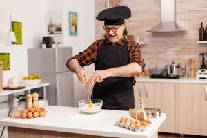 Retired chef cracking eggs for wheat flour in home kitchen. Elderly pastry chef cracking egg on glass bowl for cake recipe in kitchen, mixing by hand, kneading. photo