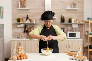 Elderly woman cracking eggs over wheat flour in home kitchen. Elderly pastry chef cracking egg on glass bowl for cake recipe in kitchen, mixing by hand, kneading photo