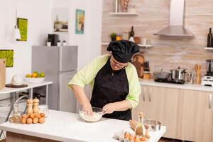 Woman mixing eggs with flour to make dough after traditional recipe on kitchen table. Retired elderly chef with uniform sprinkling, sieving sifting raw ingredients and mixing . photo