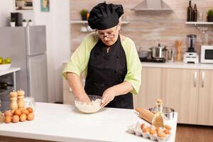 Mixing flour with eggs to make dough for tasty pasta following traditional recipe. Retired elderly chef with uniform sprinkling, sieving sifting raw ingredients and mixing . photo