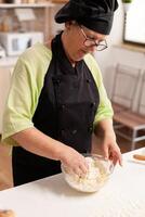Chef hand in wheat flour with eggs in home kitchen for traditional pasta. Retired elderly chef with uniform sprinkling, sieving sifting raw ingredients and mixing. photo