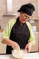 Old pastery chef wearing bonete and apron with hand in wheat flour with eggs. Retired elderly chef with uniform sprinkling, sieving sifting raw ingredients and mixing . photo