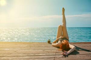 Red-haired woman is enjoying vacation at sea. Female in swimsuit is lying with raised leg on wooden pier with bottle of lemonade and sunbathing. Summer trip. Copy space for travel agency advertising. photo