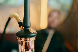 Hookah on table in lounge bar. Smoking person in blurry background. Close-up of black pipe and golden shisha flask. Leisure in oriental cafe. photo