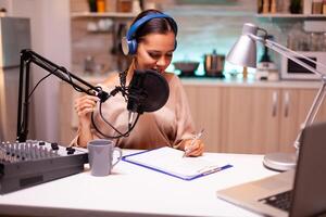 Presenter writing on clipboard and wearing headphones during podcast recording using professional microphone. Creative online show On-air production internet broadcast host streaming live content photo