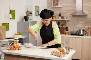 Senior woman spreading flour in home kitchen for bakery products. Happy elderly chef with uniform sprinkling, sieving sifting raw ingredients by hand baking homemade pizza. photo