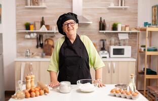 Old lady wearing chef apron and bonete in home kitchen with flour on table. Retired elderly baker in kitchen uniform preparing pastry ingredients on wooden table ready to cook homemade tasty bread. photo