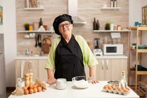 Senior woman wearing baker uniform in home kitchen smiling at camera. Retired elderly baker in kitchen uniform preparing pastry ingredients on wooden table ready to cook homemade tasty bread. photo