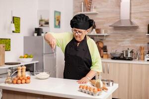 Baker spreading flour on wooden table at home in modern kitchen. Happy elderly chef with uniform sprinkling, sieving sifting raw ingredients by hand baking homemade pizza photo