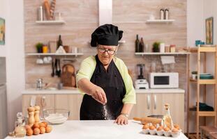 Chef sprinkling baking powder on home kitchen table wearing bonete and apron. Happy elderly chef with uniform sprinkling, sieving sifting raw ingredients by hand baking homemade pizza photo