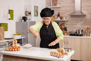 Elderly chef with uniform sprinkling flour in home kitchen wearing apron and bonette. Happy elderly chef with uniform sprinkling, sieving sifting raw ingredients by hand baking homemade pizza. photo