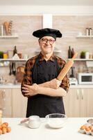 Caucasian old man wearing apron in home kitchen smiling at camera. Retired elderly baker in kitchen uniform preparing pastry ingredients on wooden table ready to cook homemade tasty bread, cakes and pasta photo