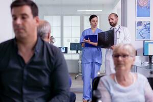 Specialist doctor holding patient x-ray explaining disease diagnosis to nurse while standing in hospital waiting area. Disabled senior woman in wheelchair waiting for medical examination photo