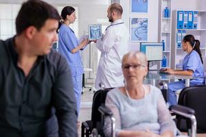 mayor mujer en silla de ruedas esperando radiografía resultados en esperando hospital zona mientras hablando con paciente. en antecedentes especialista médico examinando X rayo prueba que se discute con enfermero enfermedad tratamiento foto