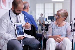 Specialist doctor showing x ray to elderly woman in wheelchair communicating test result using tablet computer. Physician explaining treatment disease during recovery while sitting in hospital area photo