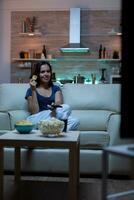 Woman enjoying the evening watching tv series at home sitting on comfortable couch dressed in pajamas. Excited amused home alone lady eating snacks and drinking juice on cozy sofa in living room. photo