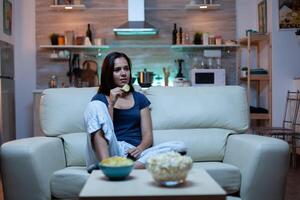 Happy woman eating popcorn on sofa and watching tv in living room at home. Excited, amused, lonely lady enjoying the evening sitting on comfortable couch dressed in pajamas in front of television. photo