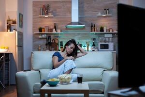 Caucasian woman falling asleep on sofa at home while watching TV. Tired exhausted lonely sleepy housewife in pajamas sleeping in front of television sitting on cozy couch in living room. photo