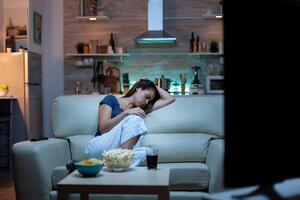 Young lady sleeping on couch watching tv. Tired exhausted lonely sleepy woman in pajamas falling asleep on sofa in front television, closing eyes while watching movie at night in living room photo