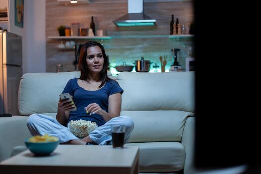Young lady relaxing at night using phone and watching tv sitting on sofa. Lonely amused happy woman reading, writing, searching, browsing on smartphone laughing amusing using technology internet photo