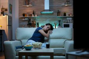 Woman falling asleep on sofa in front TV. Tired exhausted lonely sleepy lady in pajamas sleeping in front of television sitting on cozy couch in living room, closing eyes while watching movie at night photo