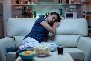 Portrait of woman sleeping on couch in living in front of television. Tired exhausted lonely sleepy lady in pajamas falling asleep sitting on cozy sofa, closing eyes while watching movie at night. photo