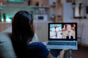 Woman in pijamas lying on sofa using laptop talking about sale report in video conference with team. Remote worker having online meeting consulting with colleagues using videocall and webcam chat photo