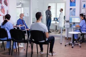 Crowded hospital waiting room with patients sitting on chairs respecting social distencing and nurse explaining treatment to elderly woman wearing face mask against covid-19. Doctor consultating man photo