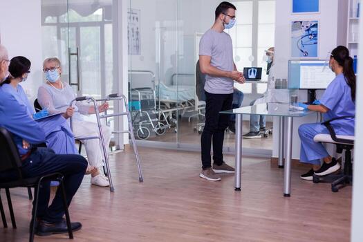 Nusre with visor against coronavirus and young patient talking in reception area, checking appointment informations. Senior disabled woman with walking frame discussing with assistant in waiting room. photo