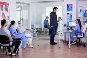 Assistant and disabled woman with walking frame talking in waiting area respecting social distancing and wearing protection mask against coronavirus. Young man visiting doctor during global pandemic photo