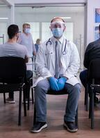 Exhausted doctor in hospital waiting room wearing surgical face mask against coronavirus looking at camera. Medic sitting in hospital hallway with medical visor protection epidemic against covid-19. photo