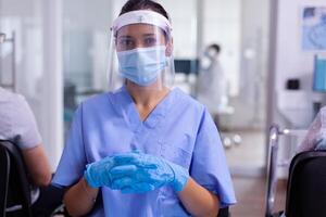 Medical assistant looking at camera wearing protective mask and visor against coronavirus sitting in hospital waiting area. Patients waiting doctor examination respecting social distancing on chairs photo