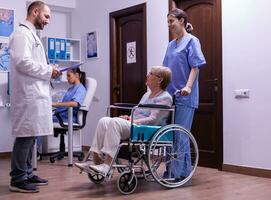 Specialist medic discussing diagnosis and recovery treatment with disabled senior woman in wheelchair. Young man doctor in hospital waiting area talking with paralyzed elderly patient photo