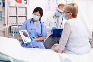 Nurse explaining heart diagnosis in cardiology to senior woman on tablet pc in time of covid pandemic. Medical assistant having a conversation with elder patient during coronavirus outbreak. photo