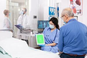 Patient looking at tablet with green screen during consultation in hospital room with nurse wearing face mask against coronavirus as safety precaution. Medical examination for infections, disease . photo