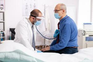 Doctor examining patient lungs using stethoscope wearing face mask as safety precaution in time of covid19. Medical practitioner wearing face mask consulting senior man in examination room during coronavirus. photo