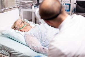 Senior woman sitting on clinic bed with oxygen mask in time of covid19. Patient in hospital room laying on medical bed with infected lung during coronavirus pandemic having respiratory problem. photo