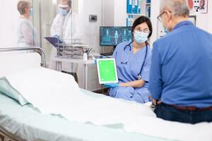 Nurse with face mask for protection against covid 19 holding tablet with green mockup while consulting senior man. Medical examination for infections, disease and diagnosis. photo